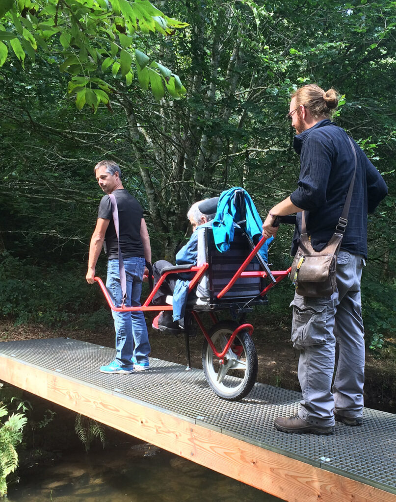 presentation_fdv_dineault deux hommes transportent un adulte en situation de handicap dans un chemin de forêt