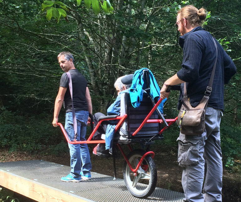 deux hommes transportent un adulte en situation de handicap dans un chemin de forêt